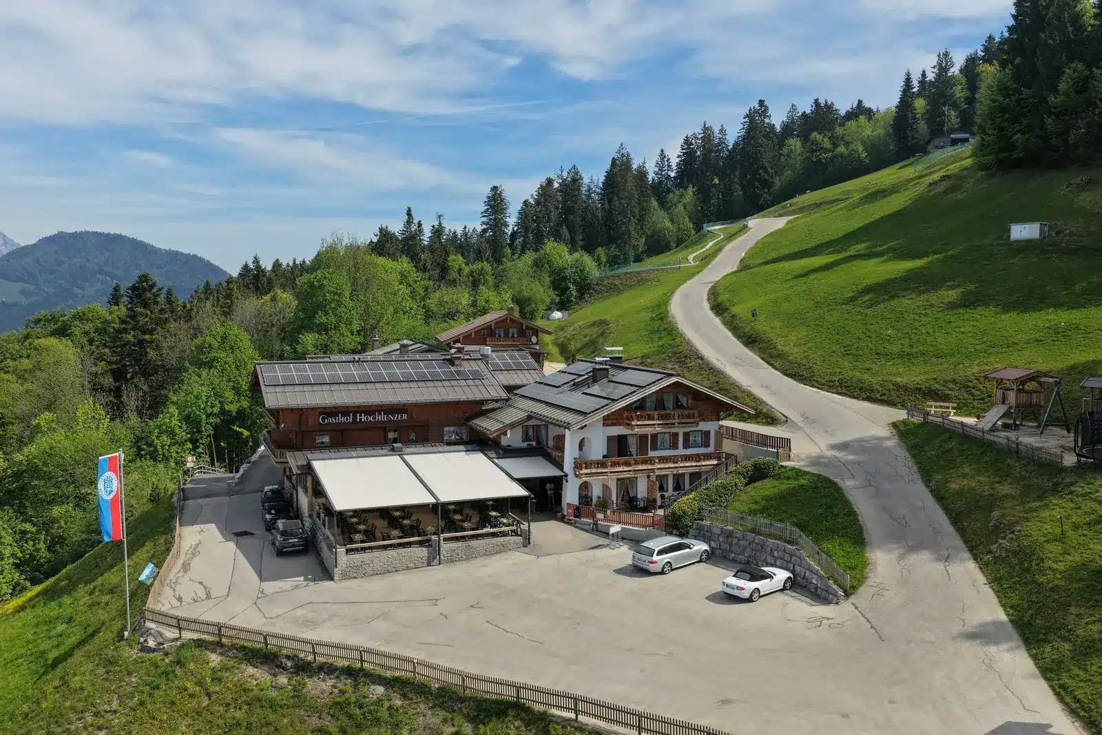 Luftbild: der Alpengasthof Hochlenzer mit Terrasse, dahinter die Almwiese und der Wald am Obersalzberg