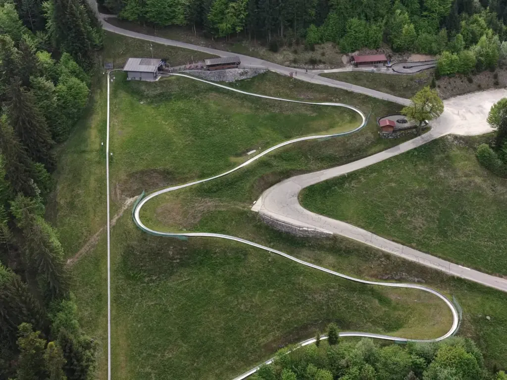 Die Sommerrodelbahn zieht sich in Kurven durch die Wiese oberhalb des Gasthofs, Luftbild